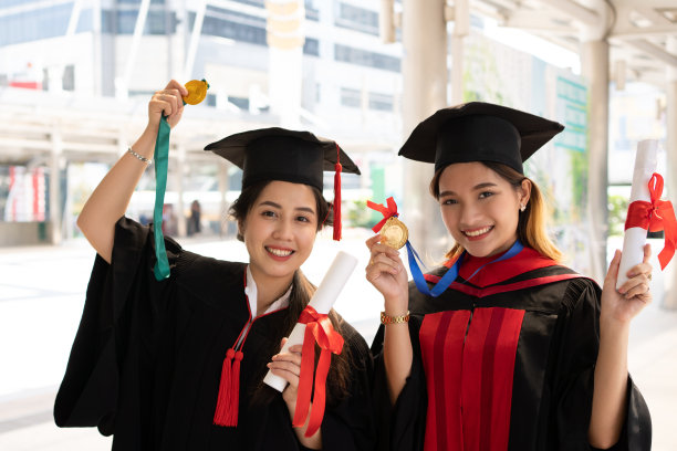两名身穿毕业礼服的亚洲女性正期待着镜头，她们手里拿着证书，面带微笑，另一个手拿着奖牌。图片下载