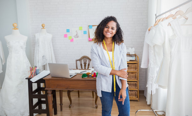 美丽的年轻非洲裔美国黑人女设计师的肖像工作使用电脑和微笑，而站在裁缝织物时装小商车间。年轻业主企业家创意女孩纺织经营理念图片下载