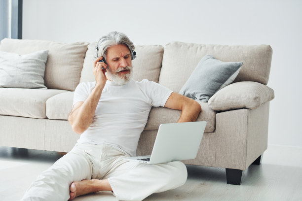 坐在沙发附近的地上，用笔记本电脑听音乐。室内有灰色头发和胡子的高级时尚现代男士图片下载