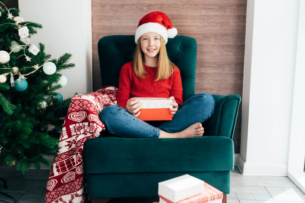 快乐微笑的小女孩戴着圣诞帽坐在家里的椅子上，拿着圣诞礼物。图片下载