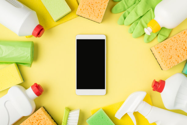 顶部视图照片的智能手机在中间洗涤剂喷雾和凝胶瓶黄色和绿色海绵粘胶布垃圾袋刷和橡胶手套孤立的黄色背景与copyspace图片下载