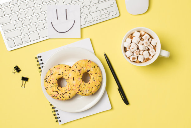 顶视图照片的工作场所贴纸笔记与画微笑的脸在键盘鼠标板与两个甜甜圈在记事本，杯子饮料与棉花糖夹夹笔孤立的淡黄色背景图片下载
