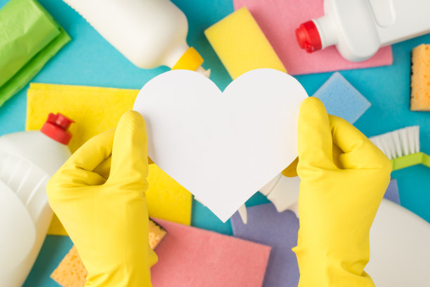 顶视图照片手在黄色橡胶手套持有白色纸心，多色海绵，粘胶布，垃圾袋，刷子和洗涤剂瓶子在孤立的淡蓝色背景与copyspace图片下载