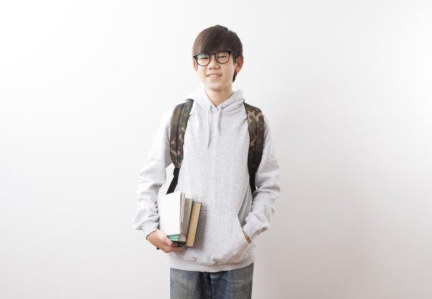 亚洲少年男生背着书包，站在白色背景下看书。图片下载