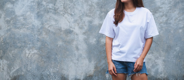 一名身穿白色t恤和蓝色牛仔短裤的年轻女子站在水泥墙背景前图片下载