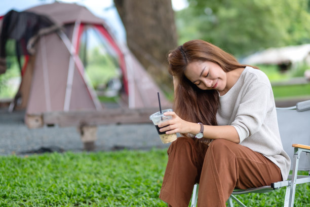 一个美丽的年轻亚洲女人的肖像，坐在露营椅与帐篷在公园喝咖啡图片下载