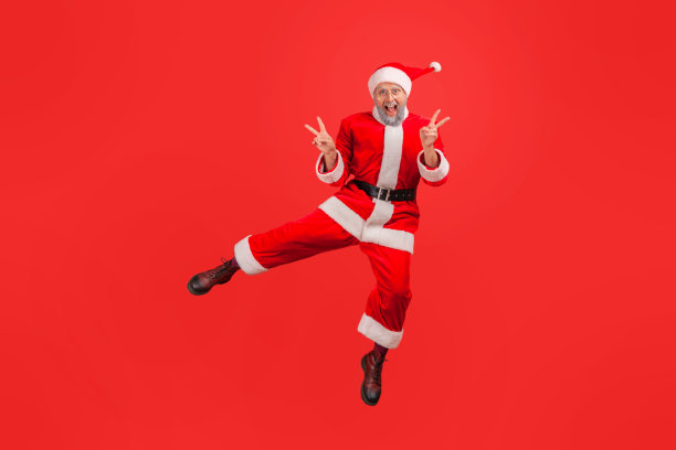 穿着圣诞老人服装的灰色胡子的快乐老人的全身肖像跳跃用手指做v的手势，庆祝冬季假期。图片下载