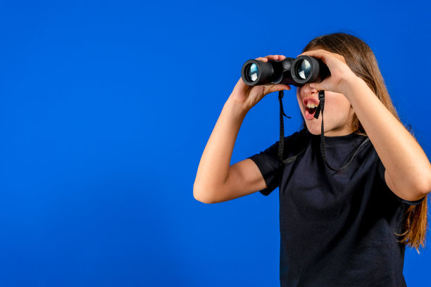 一个带着双筒望远镜的青春期前的漂亮女孩惊讶地看着她，因为她在远处发现了一些东西，在蓝色摄影棚的背景下。探索概念图片下载
