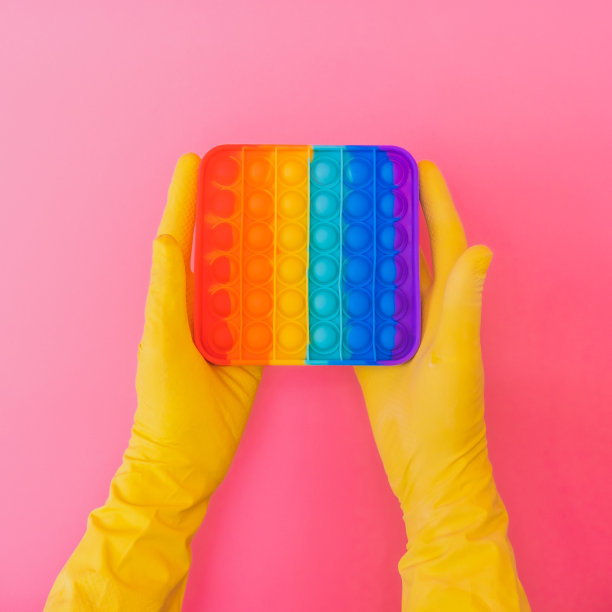 两只手在黄色橡胶手套持有彩虹抗压力pop它玩具在粉红色的背景。创造性的概念。平铺，俯视图。图片下载
