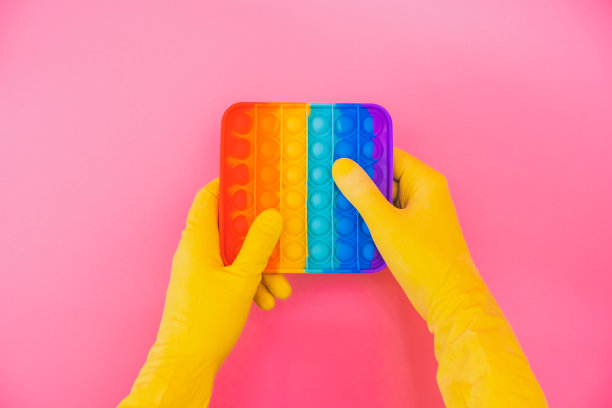 两只手在黄色橡胶手套持有彩虹抗压力pop它玩具在粉红色的背景。创造性的概念。最小的抽象艺术图片下载