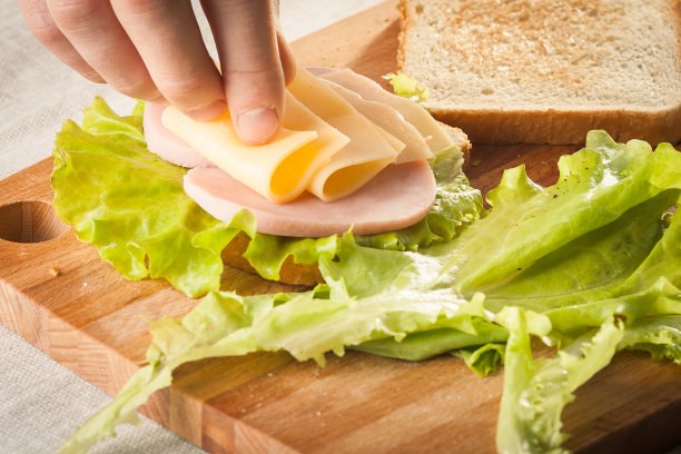 双手放在木制砧板上，用面包、奶酪、沙拉和火腿制作三明治图片下载