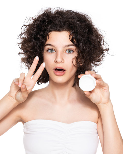 少年护肤品。美丽的少女，一头卷发，手里拿着保湿面霜，看着镜头。摄影棚里的白色背景。美容、护肤和青春期概念。图片下载