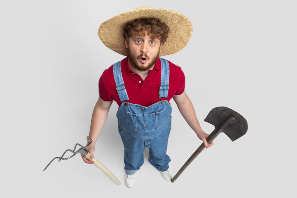 红发大胡子男人，戴草帽的农民与花园设备隔离在白色的工作室背景。职业、工作的概念。图片下载