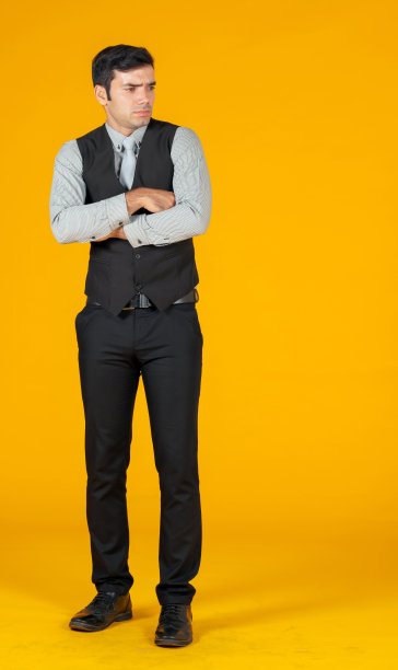 身穿灰色衬衫和黑色背心的白人商人双手交叉在胸前，惊讶地看着左边。人像在黄色背景与工作室灯光。图片下载