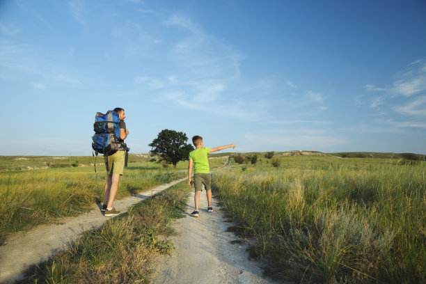 一位年轻的父亲背着背包，带着小儿子在田野和山丘间穿行，男孩站在大路上，用手指着远处的天空，看着什么东西图片下载