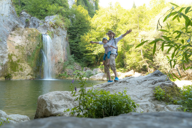 父亲和儿子一起散步，他抱着儿子，张开双臂，在瀑布前摆姿势。图片下载