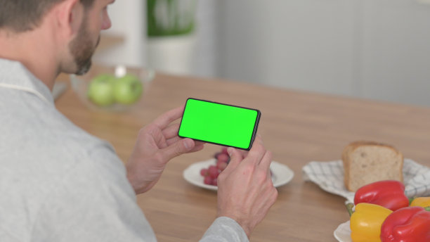 一名男子在厨房里看着带有色度键屏幕的智能手机图片下载