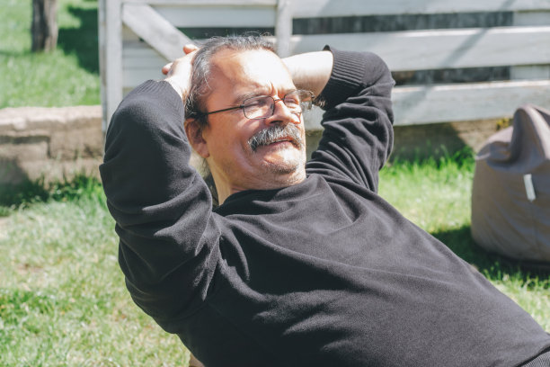 头发灰白的成年男人的肖像，戴着眼镜和胡子，坐在豆袋上，微笑着，双手放在脑后图片下载