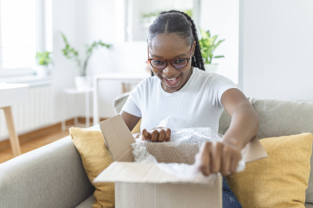 快乐的年轻女子坐在客厅沙发上打开纸箱在网上买东西，微笑兴奋的千禧女孩打开纸箱包裹订购，网上购物，好送货概念图片下载