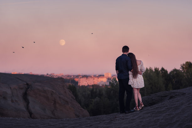 相爱的情侣相拥在一起，看着夕阳下的月亮和城市图片下载
