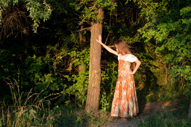 一个穿着彩色长背心裙的女孩站在森林里，手靠在一棵树上。图片下载