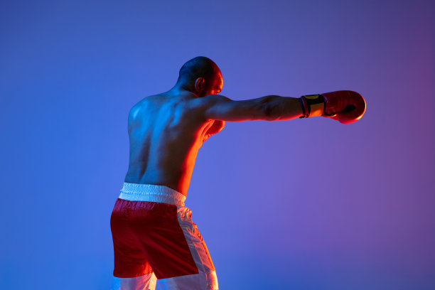 一个穿着红色短裤的职业拳击手在霓虹灯下的紫色背景下训练。后视图图片下载