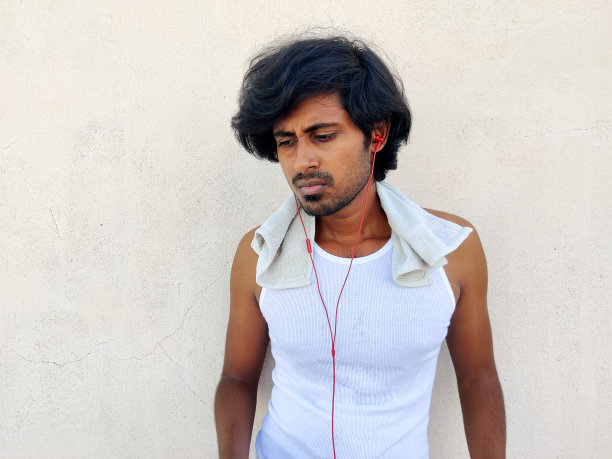 身穿白色运动服的英俊的印度年轻人正在通过耳机听音乐。白色背景图片下载