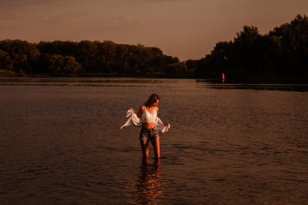 日落时一个女人在水里的照片。一个美丽的金发女郎，穿着白色的衣服和牛仔短裤，站在河边，在风景的背景下看水的侧面图片下载