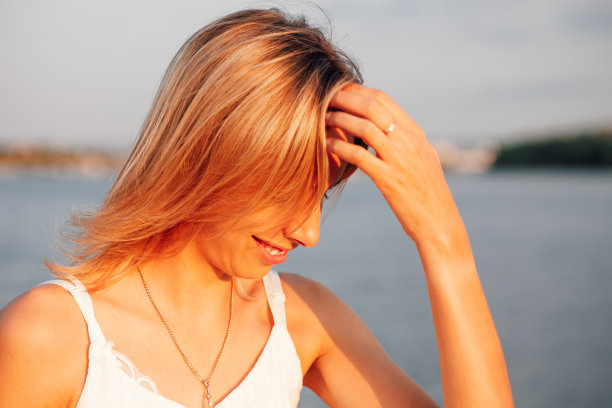 一个年轻的金发女郎的肖像在水的背景。特写照片中，一个美丽的微笑的女人穿着白色的衣服，戴着金色的珠宝，抚摸着她的头发图片下载