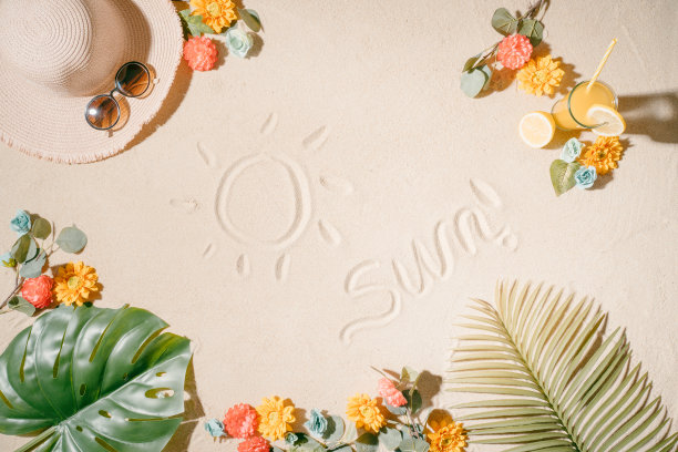 “太阳”这个词和一张画在沙滩上的太阳的图片，背景是一顶帽子，大大的太阳镜，柠檬汁，五颜六色的花，椰子和棕榈叶。暑假和假期的概念。副本的空间。图片下载