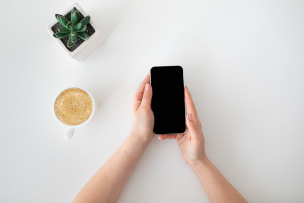 女人拿着并使用带有黑屏的现代智能手机，坐在带咖啡杯的桌子旁，俯视图图片下载