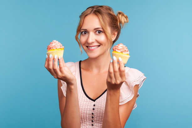 快乐微笑的年轻成年女士穿着衣服，带着两个发髻展示两个美味的蛋糕，乐观地看着镜头，提供美味的甜点。图片下载