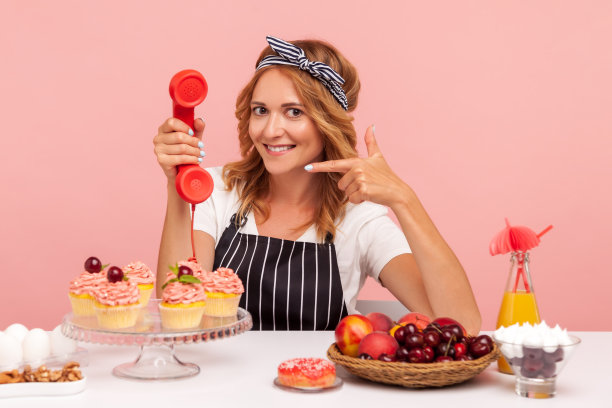 照片中的金发女人穿着白色t恤和围裙，坐在桌边，周围是糕点和水果，拿着手机，用手势叫我。图片下载