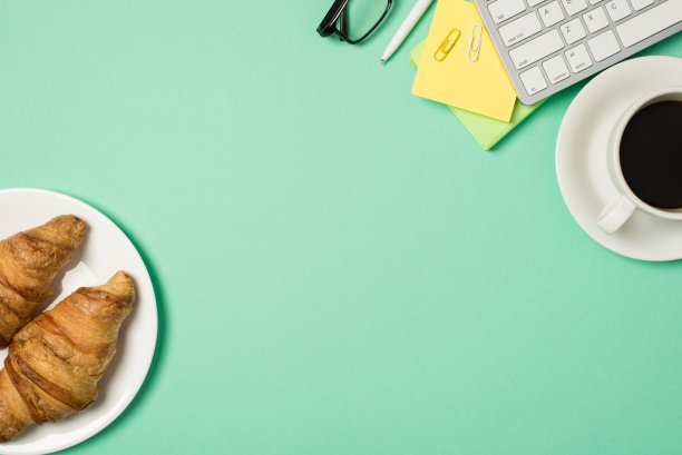 顶视图照片的工作场所键盘，便利贴，回形针，钢笔眼镜，牛角面包在盘子和咖啡杯在碟子上孤立的绿松石背景与copyspace图片下载