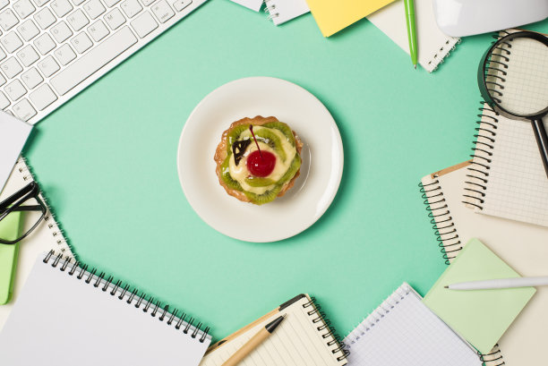 头顶的照片键盘，电脑，鼠标，放大镜，钢笔，眼镜，水果蛋糕在中间，盘子和笔记本的堆孤立在蓝绿色的背景图片下载
