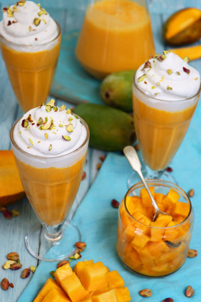 近景:芒果奶昔，上面有搅打过的奶油和切碎的开心果，还有玻璃罐切碎的芒果片，勺子，壶，芒果奶昔，绿松石木纹表面，绿松石蓝色背景图片下载