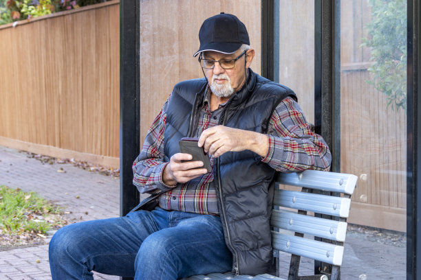 一位老人坐在郊区公交车站的候车亭里用手机查看时刻表图片下载