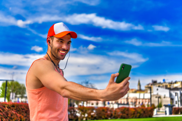 英俊的运动男在一个温暖的阳光明媚的日子里，一边走在城市里一边自拍，一边使用智能手机。图片下载