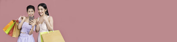 两个美丽的亚洲女人的肖像穿着裙子拿着购物袋智能手机孤立在粉红色的背景。女孩在购物，快乐女人拿着购物袋，消费主义，购物狂，生活理念的旗帜图片下载