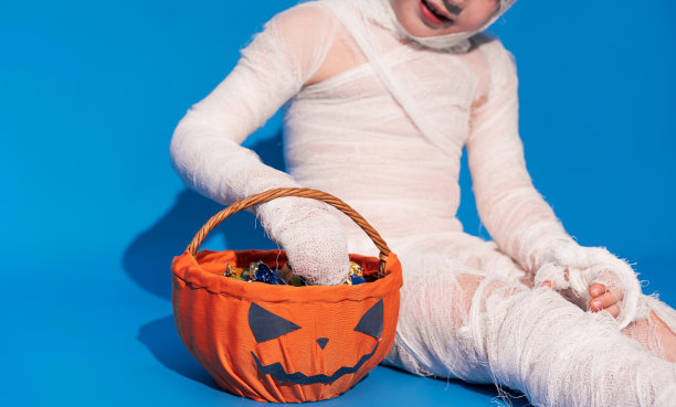 穿着木乃伊服装的孩子在蓝色背景下吃着南瓜篮子里的糖果图片下载