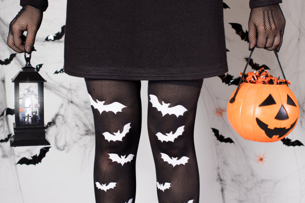 一个万圣节女孩穿着女巫服装，穿着蝙蝠袜，拿着一个发光的灯笼和一个装满糖果的南瓜桶，白色的背景上有蜘蛛网。图片下载