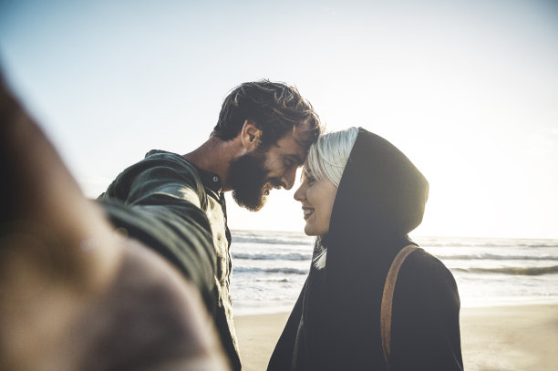 夕阳西下，一对相爱的旅行情侣在荒芜的海滩上自拍——有着迷人目光的女人和她的潮男男友在自拍时凝视着对方的眼睛图片下载