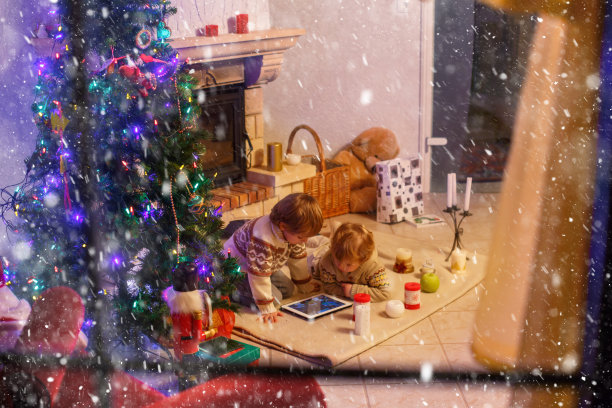 圣诞节的时候，两个兄弟姐妹带着一份惊喜礼物坐在家里的壁炉旁。孩子们，可爱的金发幼童在玩新平板电脑。从外面透过窗户。图片下载