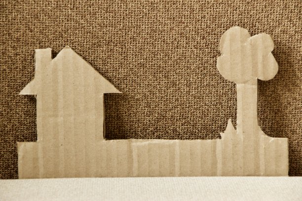 在房屋或房屋问题的概念下，将棕色纸板切割成房子的形状，烟囱树和草丛，编织棕色沙发作为背景图片下载