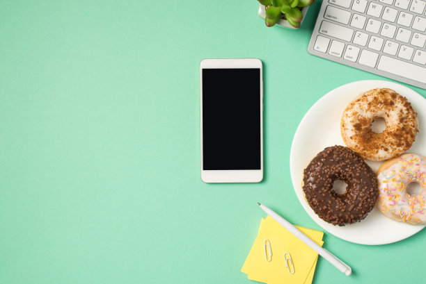 顶视图照片的工作场所键盘，笔夹，便签纸plant三个甜甜圈在盘子和智能手机在孤立的蓝绿色背景空白图片下载