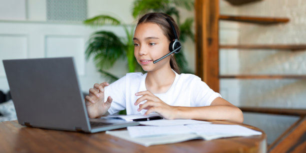 一个女孩戴着耳机在网上上课的照片。她坐在笔记本电脑显示器前，在线做作业，并向老师咨询图片下载
