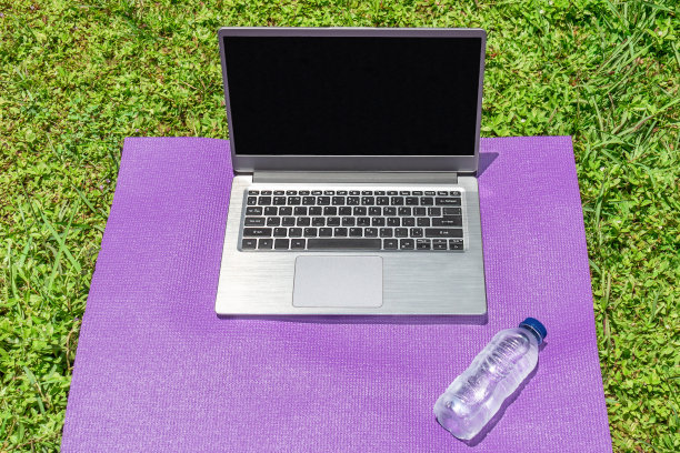 一台笔记本电脑和一瓶水放在瑜伽垫上，背景是绿色的草地。在线健身图片下载