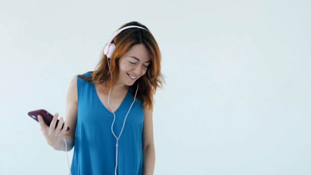 积极微笑的年轻红发女子，戴着粉色耳机，身穿蓝色连衣裙，一边听音乐一边微笑。她独自一人在工作室白色的墙上欣赏着歌舞。情感的女性肖像图片下载