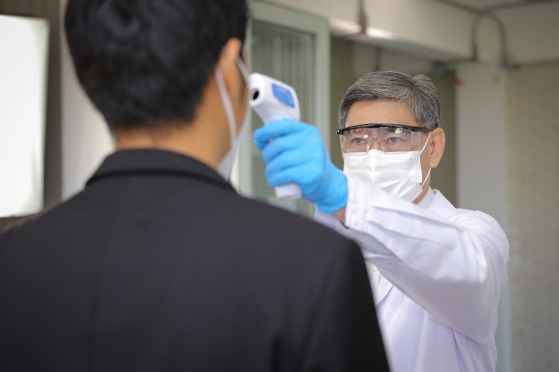 身穿个人防护服的工作人员正在用红外线温度计测量穿着西装的人的体温。图片下载