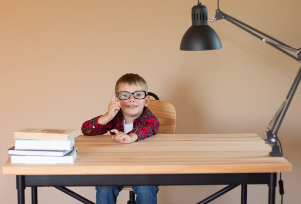 一个戴着眼镜的快乐小男孩坐在桌旁，用手指着什么东西。回到学校的概念。Copyspace图片下载